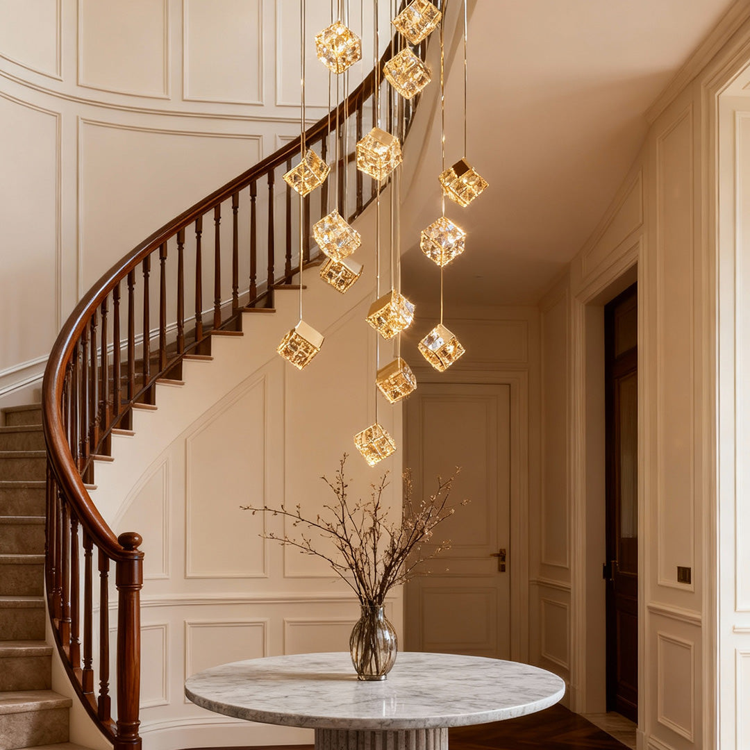 Golden crystal cubes hanging at staggered heights beside a curved staircase with a round marble table and a vase of branches below