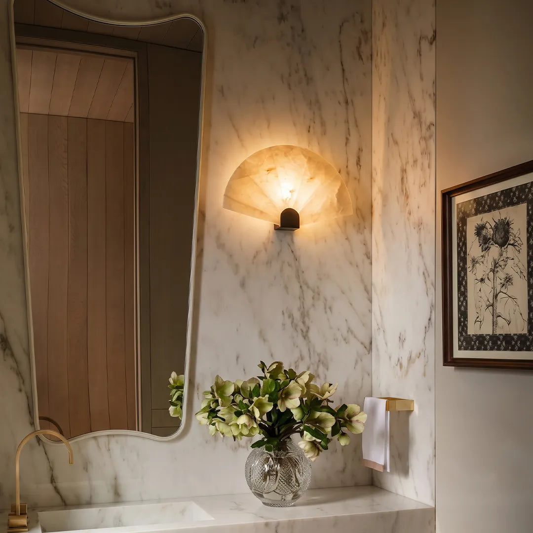 Carved marble fan sconce glowing beside a veined marble wall, a curved mirror and a vase of flowers
